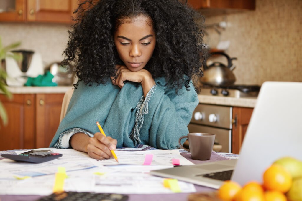 Unhappy depressed young dark skinned housewife managing domestic budget alone at night, making notes with pencil, sitting at kitchen table with open laptop, documents, calculator and cup of coffee